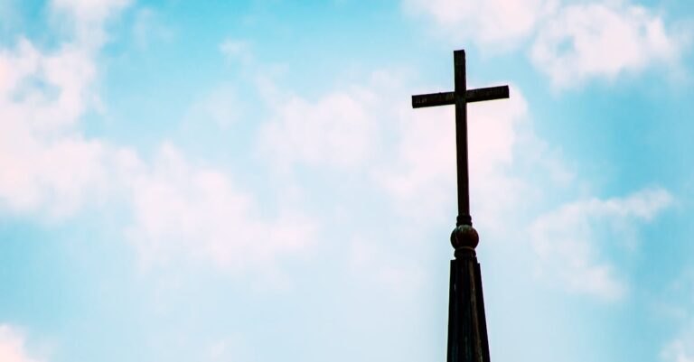A church spire with a cross silhouetted against a bright blue sky with clouds.