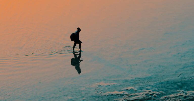 A person walks through calm water at sunset, casting a shadow as the sun sets.