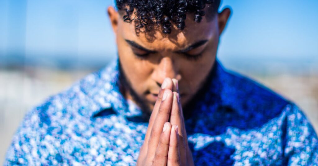 Adult man with curly hair praying outdoors under a clear blue sky, wearing a patterned shirt.